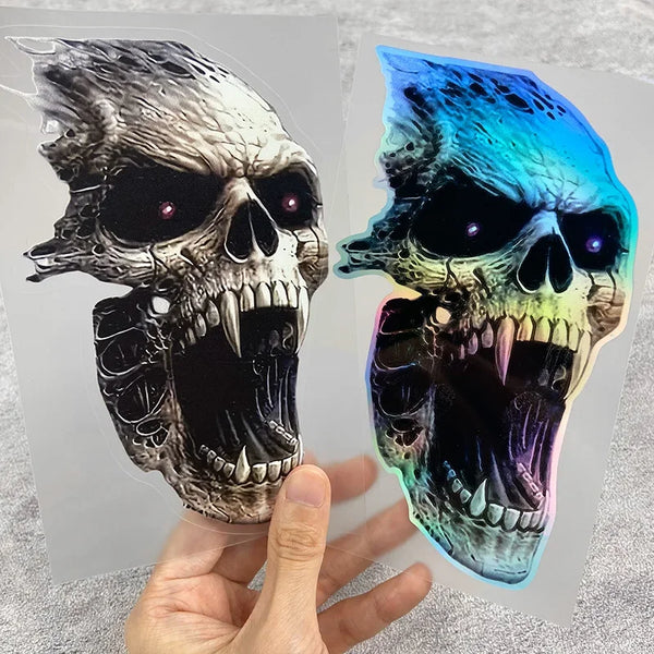 A hand holds a waterproof vinyl skull decal featuring a detailed, menacing skull with iridescent blue and green coloring.