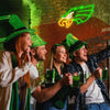 Four people cheering while holding green drinks in front of a brick wall with an illuminated neon sign.