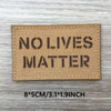 A rectangular tan woven patch with black text reading NO LIVES MATTER on a weathered wooden surface.