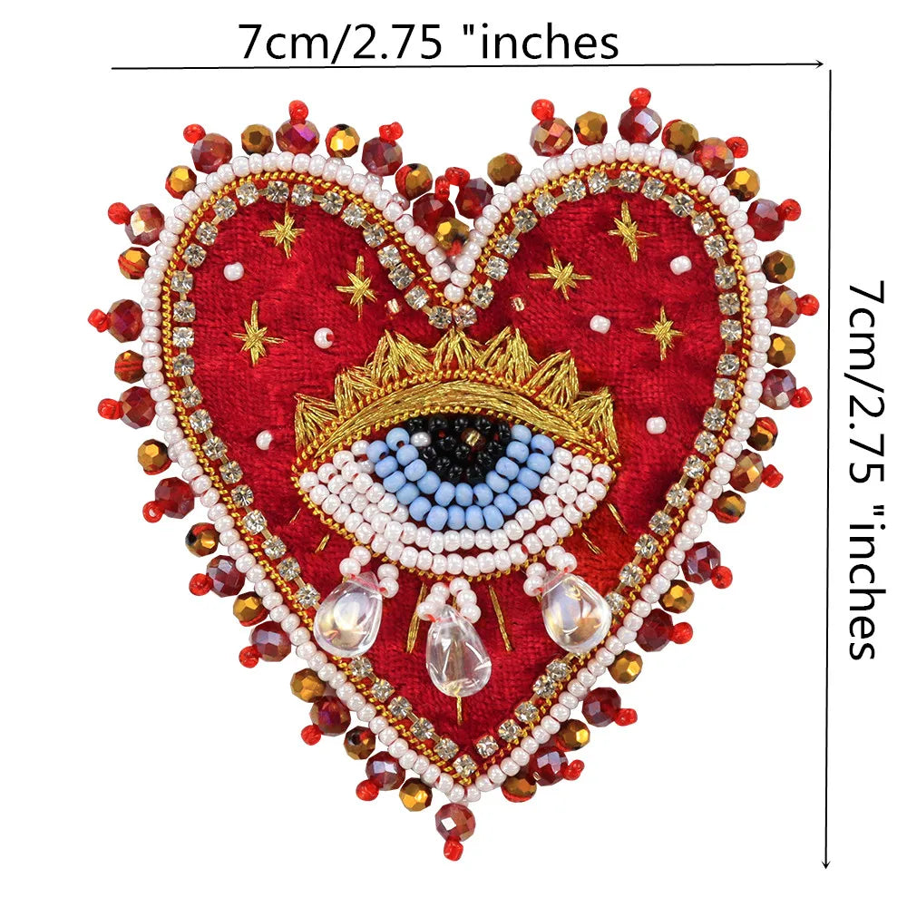 A heart-shaped patch embroidered with red fabric, gold and blue rhinestones, and a central eye design.