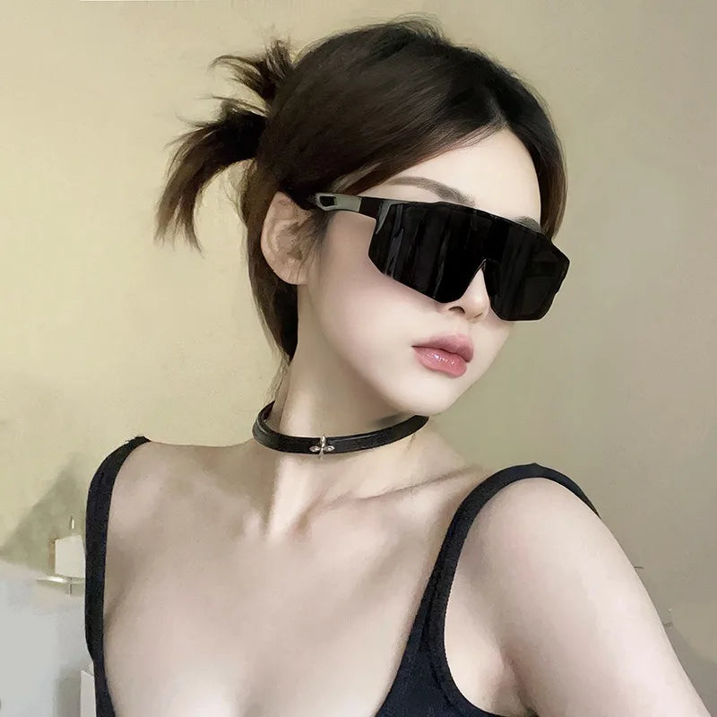 A woman wearing black sunglasses with a thin gold chain necklace against a light background.