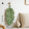 A hand towel shaped like a banana leaf hangs on a wall next to a cream-colored knitted pillow and a small white sheep.