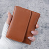 A hand holds a brown slim triple-fold PU leather wallet against a gray background.