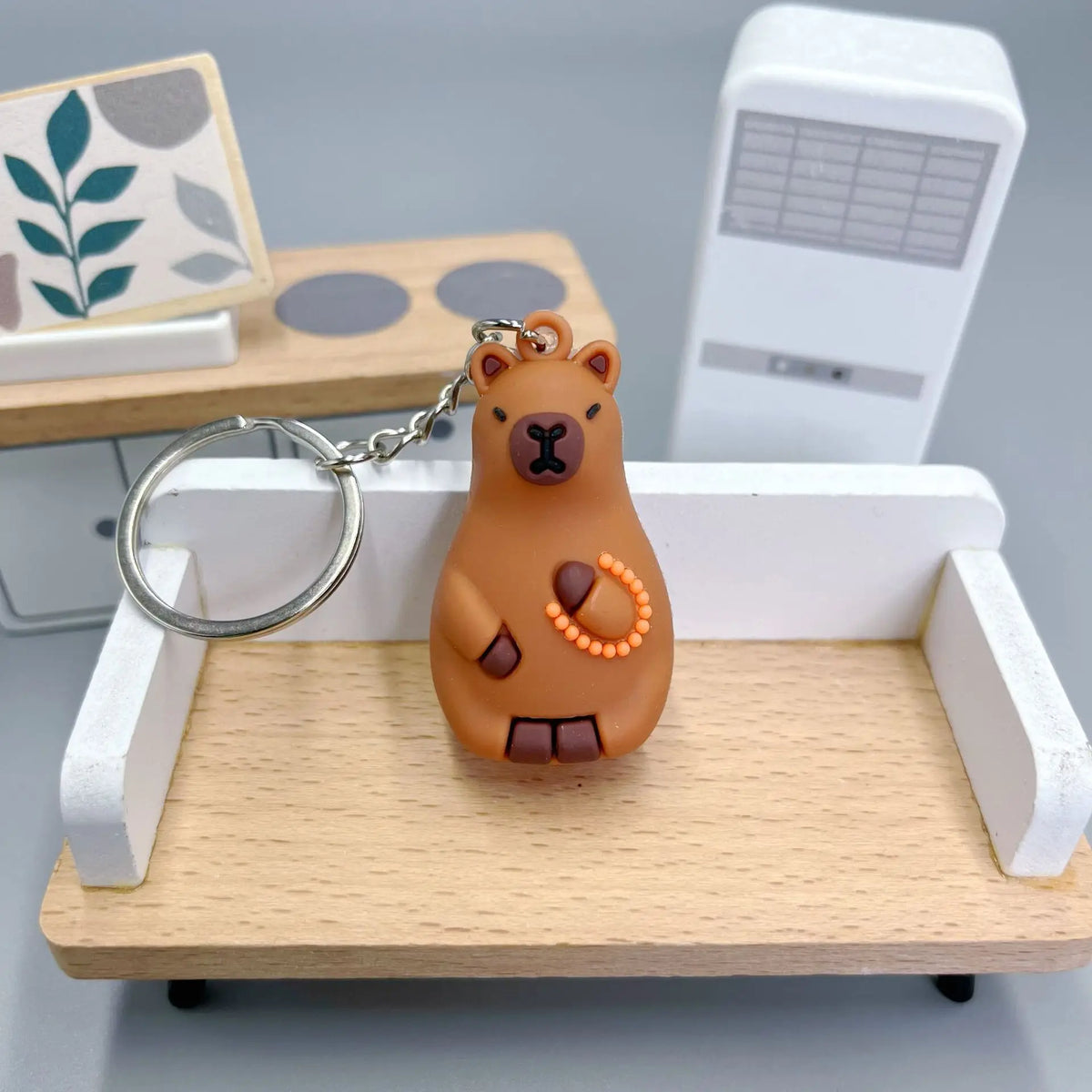 An adorable capybara keychain charm with a gold chain resting on a light wooden tray.