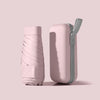 A pink compact travel umbrella is shown with its protective capsule case, featuring a gray strap.