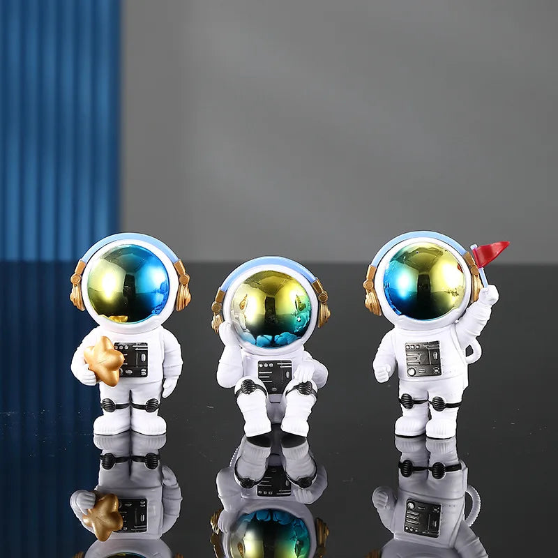 Three handcrafted astronaut figurines in white and gold resin displayed on a reflective surface.