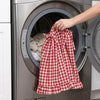 A hand is placing a red and white plaid oven skirt with a ruffled trim into the drum of a stainless steel dishwasher.