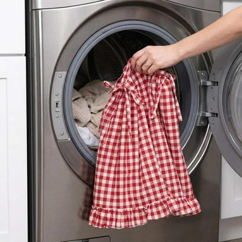 A hand is placing a red and white plaid oven skirt with a ruffled trim into the drum of a stainless steel dishwasher.