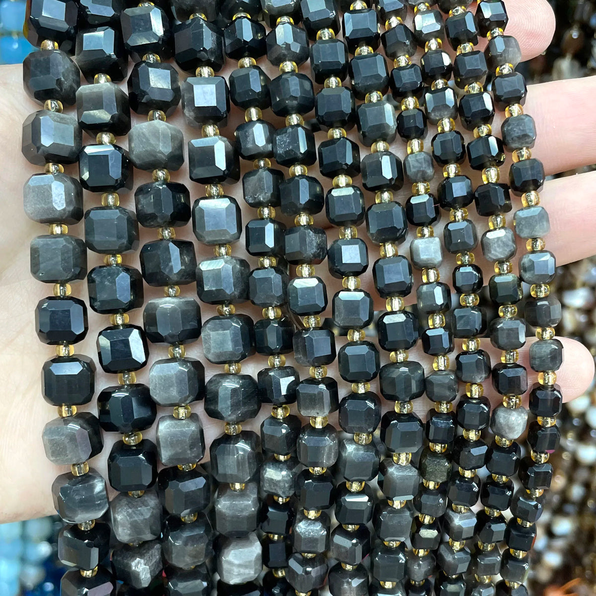 A hand holds a strand of natural stone square faceted beads in various shades of black and gray.