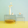 Clear acrylic display risers holding a white flower vase and a small ceramic animal figurine on a yellow surface.