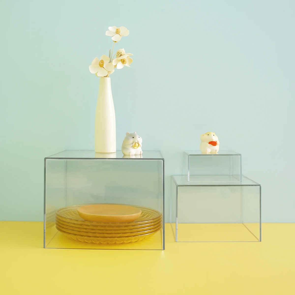 Clear acrylic display risers holding a white flower vase and a small ceramic animal figurine on a yellow surface.