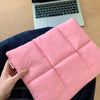Eine pinke gesteppte Puffer-Laptoptasche, von einer Hand gehalten, auf dunkelblauem Jeansstoff neben einem Laptop.