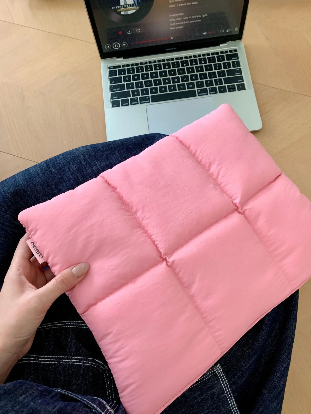 Eine pinke gesteppte Puffer-Laptoptasche, von einer Hand gehalten, auf dunkelblauem Jeansstoff neben einem Laptop.