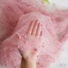 A hand is gently holding and spreading sheer pink tulle fabric adorned with small pearls.
