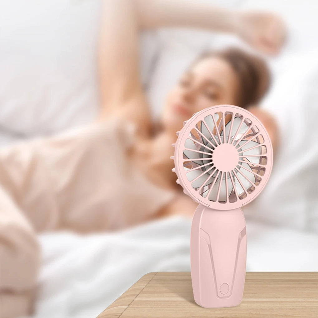 A pink handheld fan is resting on a wooden surface next to a person lying in bed.