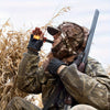 A person in camouflage is using a portable crow call whistle outdoors amidst dry grass.