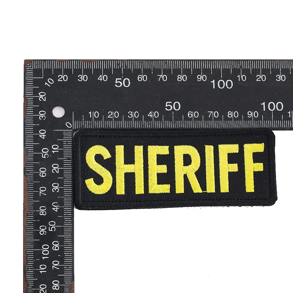 A black embroidered patch with the word SHERIFF in yellow text against a ruler scale.