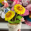 Handcrafted crochet sunflower with a green stem growing in a beige pot surrounded by colorful yarn flowers