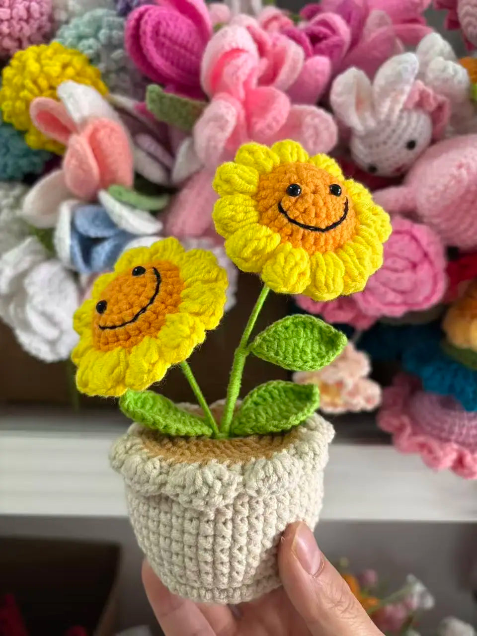 Handcrafted crochet sunflower with a green stem growing in a beige pot surrounded by colorful yarn flowers