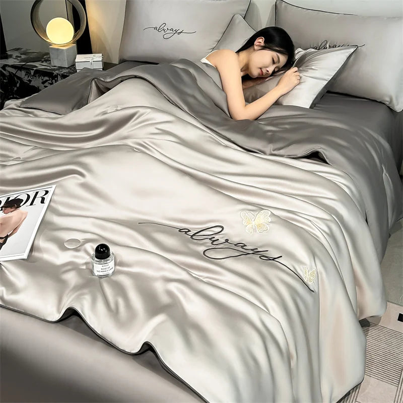 A luxurious silver comforter with embroidered 'always' text is draped over a bed, next to a lamp.