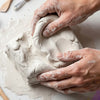 Hands sculpt a form using light beige air-dry clay on a white surface.