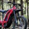 Red Sur-Ron electric dirt bike with front and rear fenders in a forest setting.