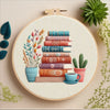 A circular embroidery kit featuring stacked books with floral designs and various colored threads on a light background.