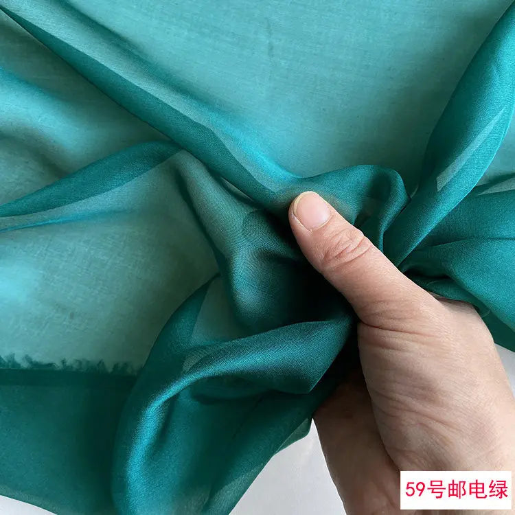 A hand is folding a piece of teal green silk chiffon fabric.