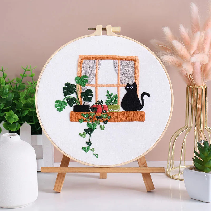 A circular embroidery kit featuring pre-printed floral and cat designs on a white canvas, displayed on a wooden easel.