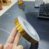 A hand holds a roll of gold-colored self-adhesive waterproof tile gap sealing tape on a gray surface.