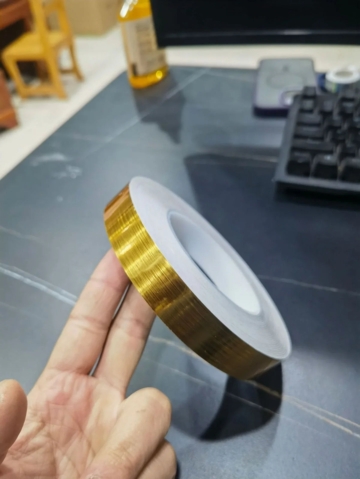 A hand holds a roll of gold-colored self-adhesive waterproof tile gap sealing tape on a gray surface.