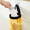A hand is pulling a white paper towel from a yellow mesh plastic bag dispenser mounted on a white wall.