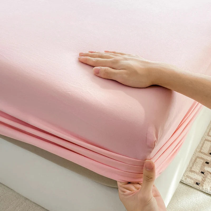 A hand is folding a pink cotton fitted sheet with an elastic band on a white bed frame.