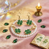 Gold and pearl clover leaf keychain charms displayed on pink silk fabric with a lit candle and green gemstones.