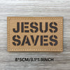 A rectangular tan woven patch with black text reading JESUS SAVES on a weathered wooden surface.