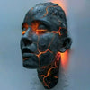 A dark, cracked face sculpture illuminated by glowing orange light mounted on a wall.
