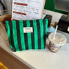 A green and black striped canvas cosmetic bag is displayed next to a cup of iced coffee.