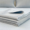 A stack of white and gray quilted linen fabric covers resting on a white bedspread.