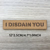 A rectangular tan patch with the text I DISDAIN YOU engraved on it, shown against a light wooden background.