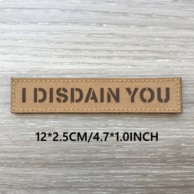 A rectangular tan patch with the text I DISDAIN YOU engraved on it, shown against a light wooden background.
