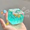 A hand holds a translucent teal ocean squishy stress ball filled with colorful sea creatures and glitter.