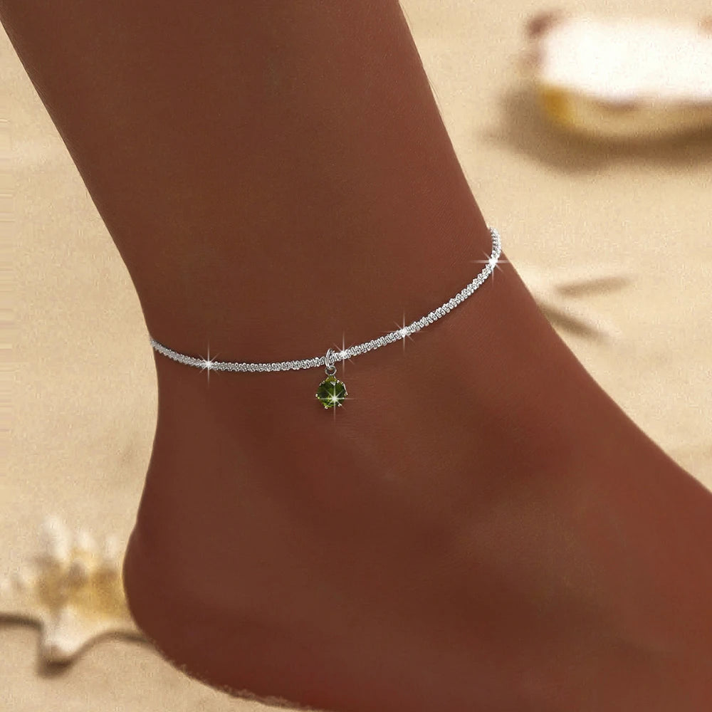 A dainty silver chain anklet with a layered design and a small green gemstone charm worn on a person's ankle.