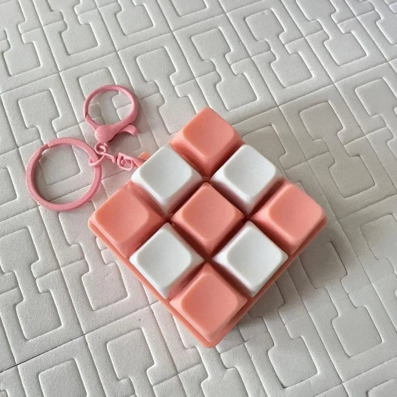 A pink tactile clicking keyboard keychain fidget toy with white square buttons resting on a white grid surface.
