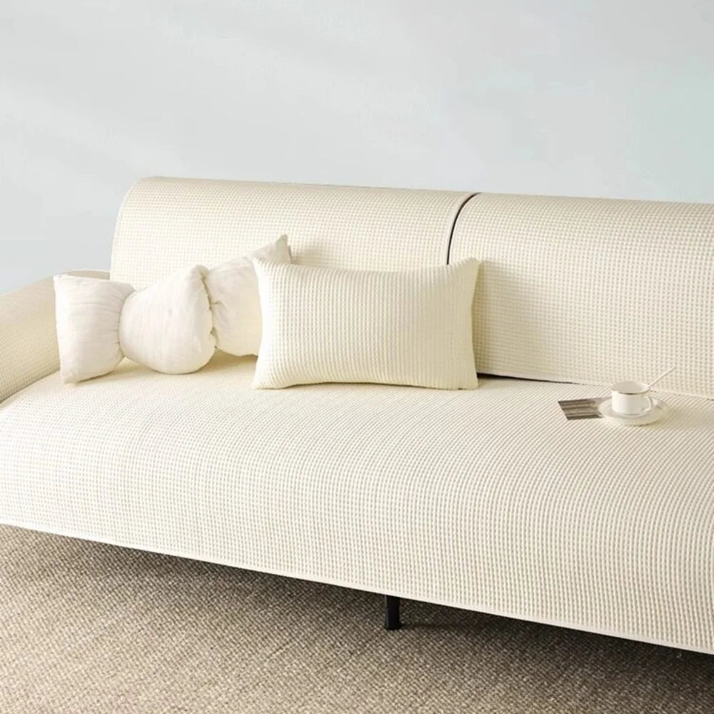 Cremelfarbener rutschfester Sommersofaüberzug mit weißen Kissen auf einem hellbeigen Sofa.
