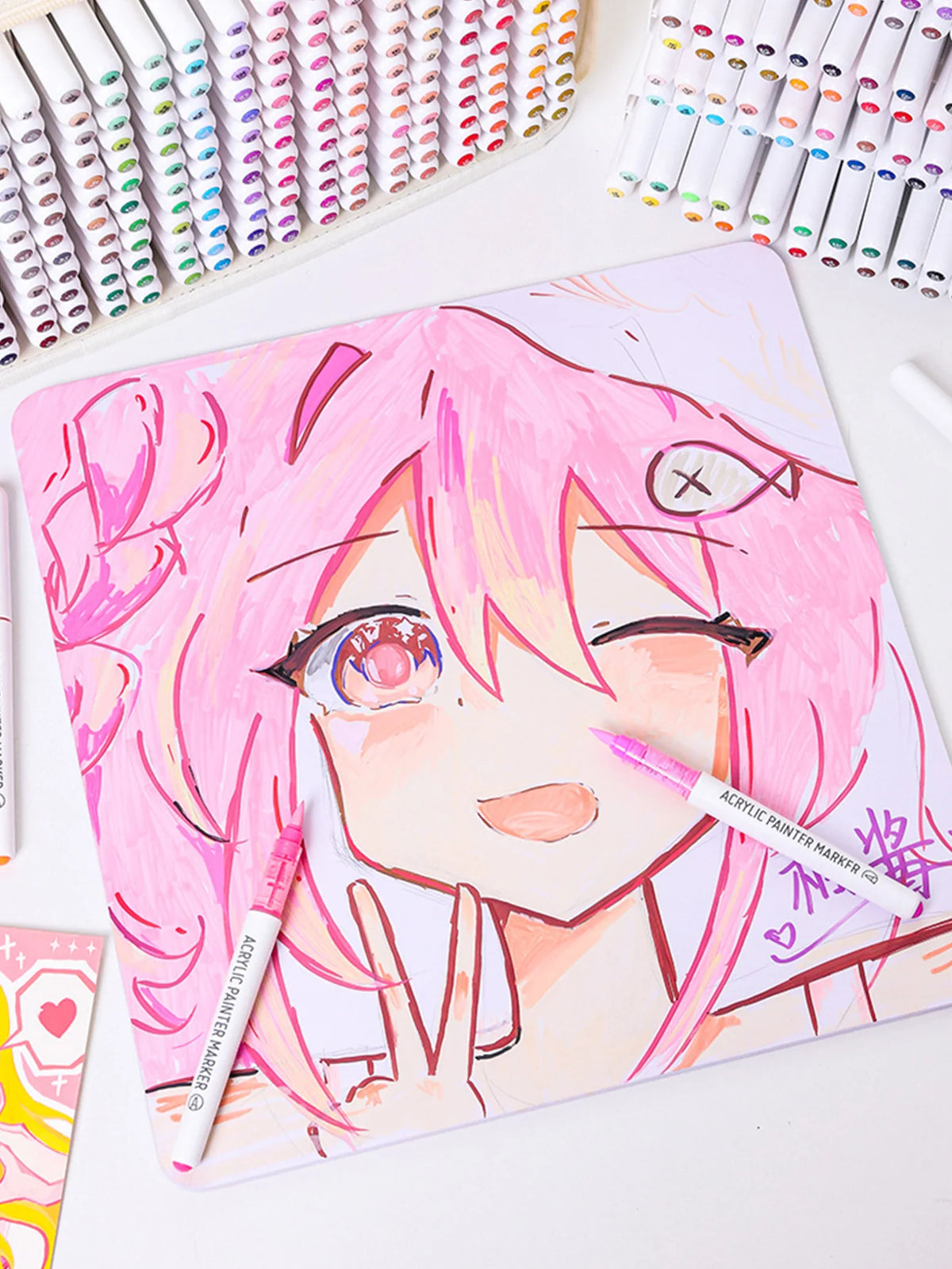 A close-up of a hand-drawn anime style face in pink and white, surrounded by acrylic paint markers.
