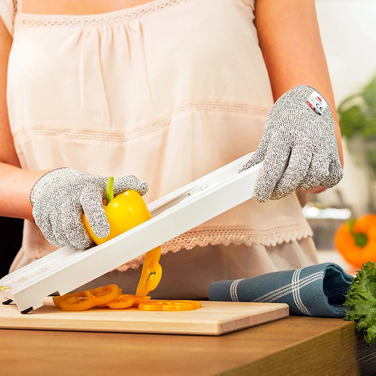 A person wearing gray cut-resistant work gloves is holding a yellow bell pepper on a white cutting board.