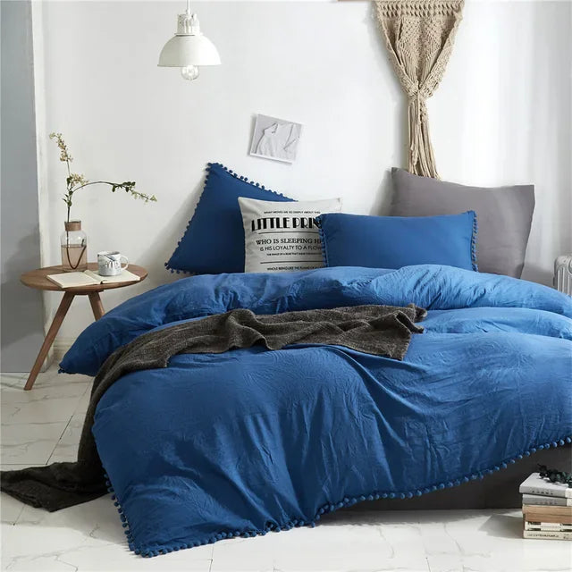 A deep blue duvet cover set with pom tassels and a gray throw blanket on a bed.