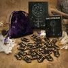 A set of wooden rune stones, a guide book, and a purple velvet bag are displayed on a rustic surface.