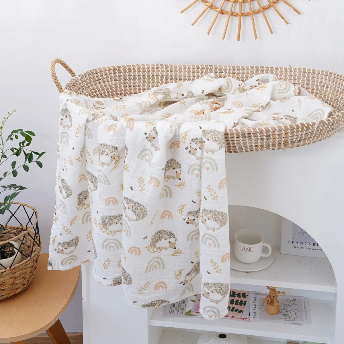 A white muslin baby swaddle blanket with a light brown and gold pattern is draped over a woven basket on a white shelf.
