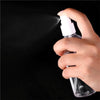 A hand is spraying a clear liquid from a white portable refillable travel spray bottle against a dark background.